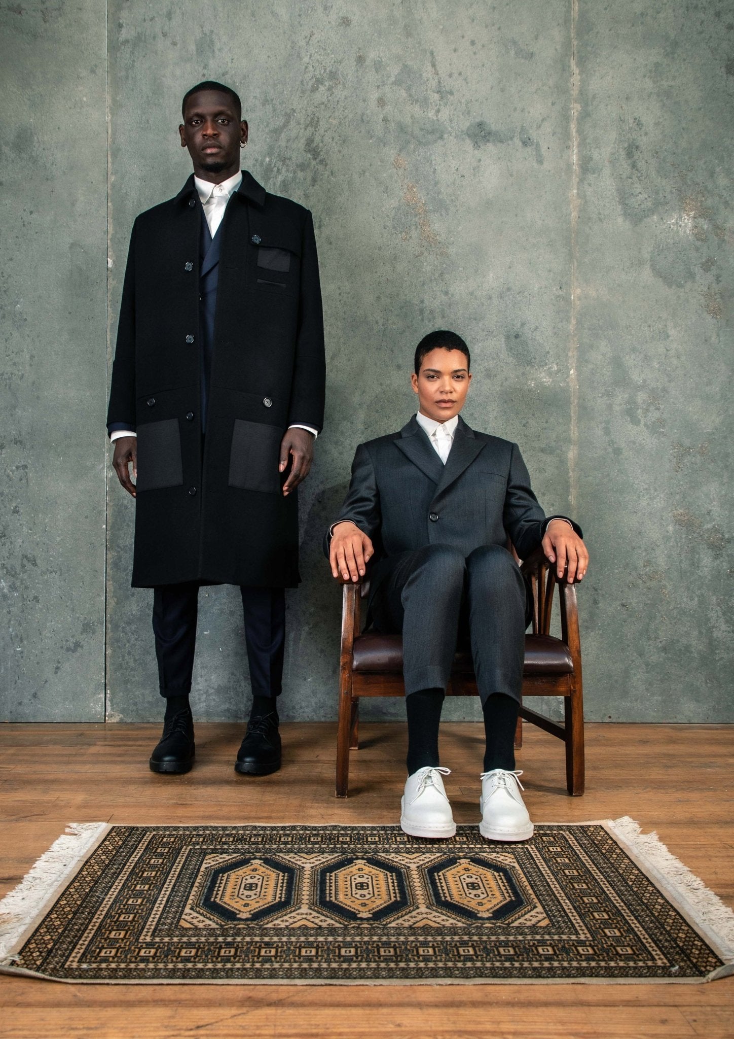 LE MANTEAU NOIR EN LAINE & CACHEMIRE porté par un homme avec une femme en costume, sur un tapis au design moderne.