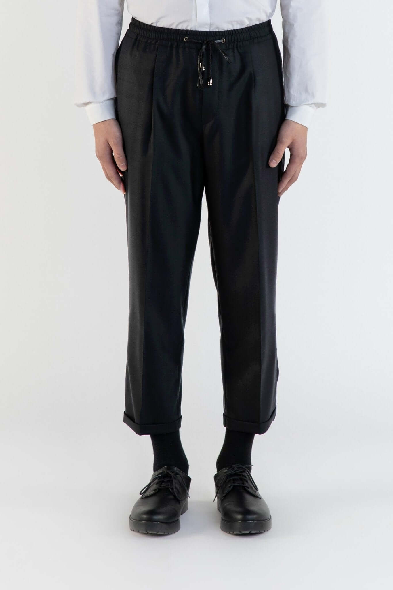 LE PANTALON NOIR À PLIS & CEINTURE ÉLASTIQUÉE EN LAINE porté par un modèle sur fond neutre.