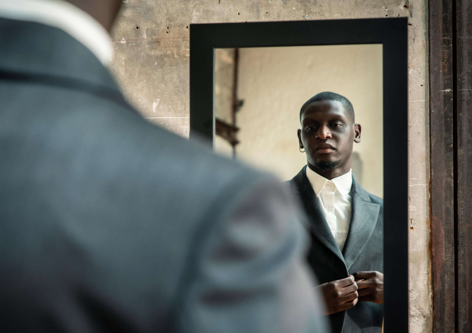 A man in a stylish suit adjusting his collar while looking in a mirror, showcasing modern fashion and confidence.